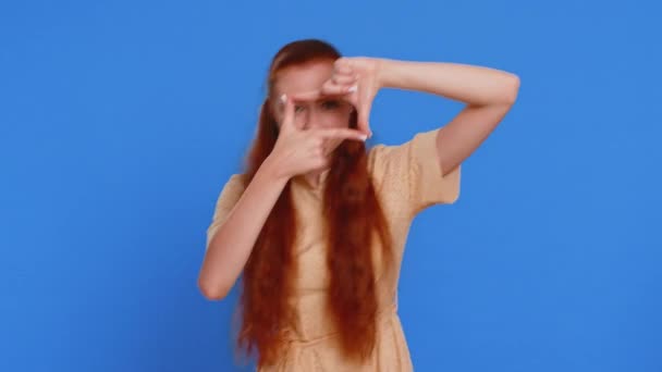 Jeune femme adulte photographe gestuelle cadre photo avec les mains, regarde à travers les doigts et en se concentrant sur le moment intéressant, imitant zoom et recadrage belle image. Fille rousse sur fond bleu - Séquence, vidéo