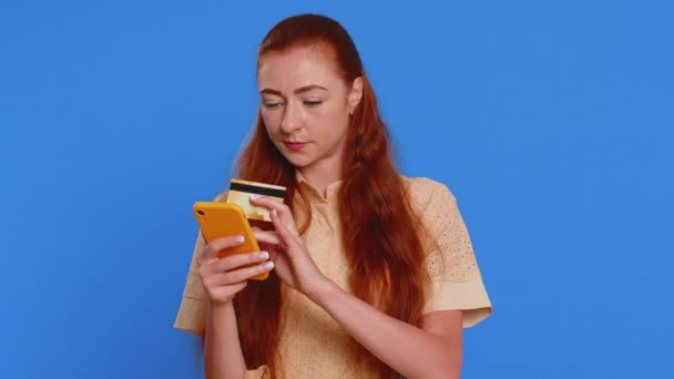 Sincere young woman customer using credit bank card and smartphone while transferring money, purchases online shopping, payment. Finance and internet. Redhead girl isolated alone on blue background - Footage, Video