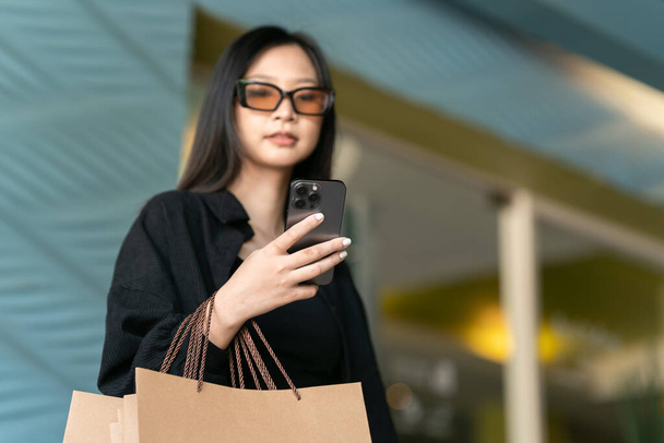 Junge Frau, die mit Einkaufstaschen am Straßenrand steht und ihr Handy benutzt. Einkäufe, schwarzer Freitag, Rabatte. - Foto, Bild