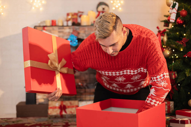 Χριστουγεννιάτικη ιδέα. όμορφος άνδρας κάθεται έκπληκτος και ανοίγοντας κουτί δώρου με φόντο χριστουγεννιάτικο δέντρο. Γιορτινή ατμόσφαιρα πάρτι. - Φωτογραφία, εικόνα