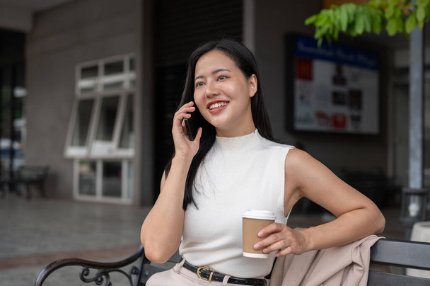 Egy vonzó és boldog Y-generációs ázsiai nő telefonál és kávét kortyolgat, miközben egy padon pihen a városban egy derűs napon. Emberek és technológiai koncepciók - Fotó, kép