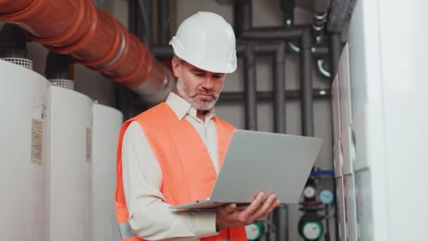 Serious bearded specialist holding wireless computer while registering data on workshop background. Qualified elderly man in white hard hat and orange vest working in field of industrial manufacture. - Footage, Video