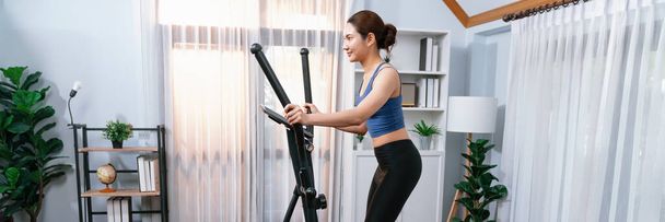 Energinen ja vahva urheilullinen aasialainen nainen juoksee elliptisellä juoksukoneella kotona. Sopivan kehon harjoittaminen ja sitoutuminen terveelliseen elämäntapaan kotiharjoittelulla ja koulutuksella. Panoraama voimakas - Valokuva, kuva