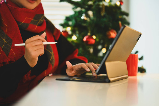 Νεαρή γυναίκα σε προειδοποιητικά ρούχα κάθεται από διακοσμημένο χριστουγεννιάτικο δέντρο και χρησιμοποιώντας ψηφιακή ταμπλέτα. - Φωτογραφία, εικόνα