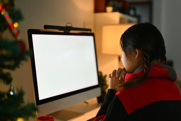 Вид сбоку молодой женщины в свитере с компьютером возле украшенной елки в уютной гостиной. - Фото, изображение