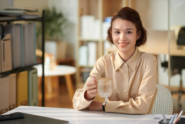 Sorrindo jovem empresária bebendo xícara de chá quando trabalhava na mesa de escritório - Foto, Imagem