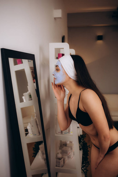 Eine Frau in schwarzer Unterwäsche mit kosmetischer Gesichtsmaske sieht sich selbst im Spiegel an - Foto, Bild