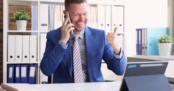 Hombre de negocios alegre hablando en el teléfono inteligente sostiene los pulgares hacia arriba y mira en el monitor de la tableta. Concepto de negocio en línea exitoso - Imágenes, Vídeo
