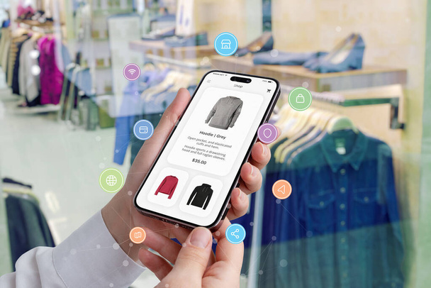 Dłonie kobiety posiadają smartfon z aplikacją zakupową otoczoną ikonami zakupów, a butik prezentuje ubrania w tle - Zdjęcie, obraz