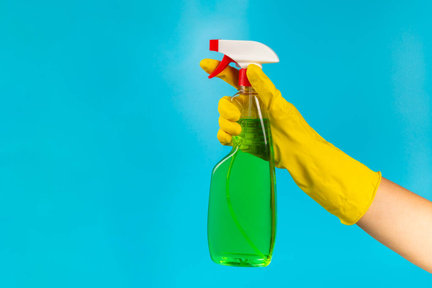 Ręka czyściciela w żółtej gumowej rękawiczce ochronnej trzymającej butelkę z chemikaliami na niebieskim tle. Firma sprzątająca. Wiosenne porządki. Miejsce na tekst lub logo. - Zdjęcie, obraz