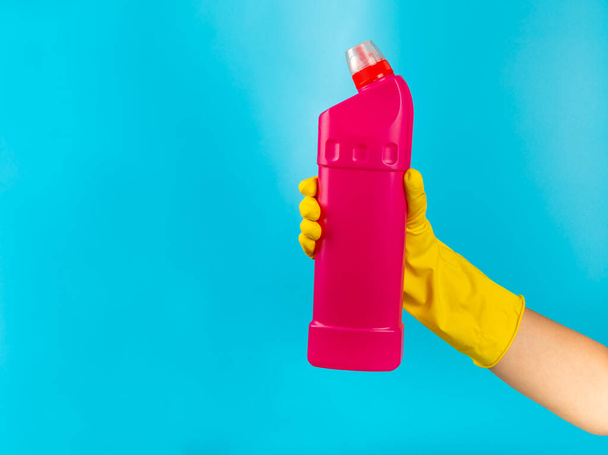 A cleaner's hand in a yellow rubber protective glove holding a bottle with a cleaning chemical on a blue background. Commercial cleaning company. Spring regular cleaning. Space for text or logo. - Photo, Image