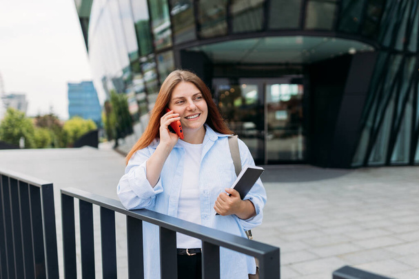Een jonge roodharige zakenvrouw die een telefoontje pleegt via een mobiele telefoon in de stad. Middelbare leeftijd vrouw aan het praten op de smartphone, wandelende stad stedelijke straat op kantoor centrum. - Foto, afbeelding