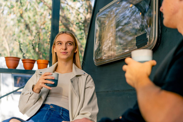 Ein junges schönes Mädchen trinkt Tee und spricht mit ihrem Freund, während sie auf der Terrasse in der Nähe eines Wohnmobils im Wohnmobil sitzt - Foto, Bild