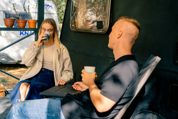 A young couple a man and a girl are drinking tea on the terrace near their motorhome in a camper - Photo, Image