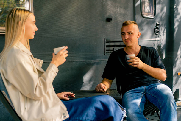 Ein junger gutaussehender Mann und ein Mädchen sitzen auf der Veranda eines Wohnmobils in einem Wohnmobil und trinken Tee und unterhalten sich begeistert. - Foto, Bild