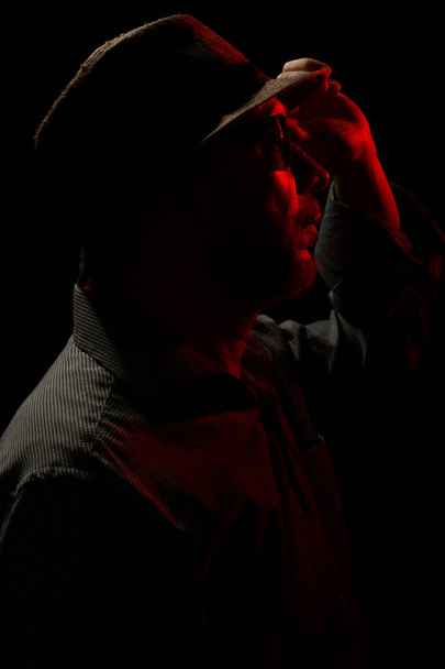 Hienovarainen muotokuva miehestä, jolla on hattu ja aurinkolasit pimeydessä ja punainen valo kasvoilla. Pimeän huoneen taide. - Valokuva, kuva