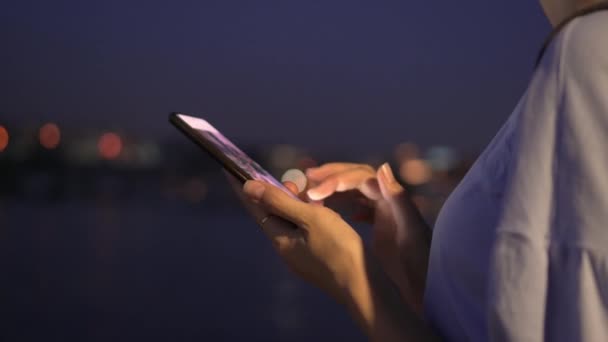 Close-up of woman's hands holding marton while standing on Night City street. Portrait of woman using mobile phone on background of night city lights. Caucasian blonde looking at smartphone screen. - Footage, Video