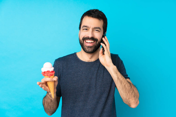 Joven con un helado de corneta sobre fondo azul aislado manteniendo una conversación con el teléfono móvil con alguien - Foto, imagen
