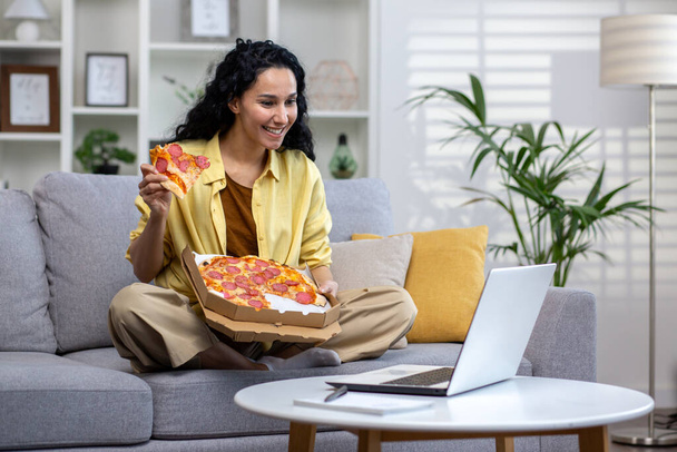 Vidám és boldog nő eszik pizza egyedül otthon, spanyol nő elégedett az időben történő élelmiszer-szállítás, a laptop nézni online video stream, ül a kanapén a nappaliban otthon. - Fotó, kép
