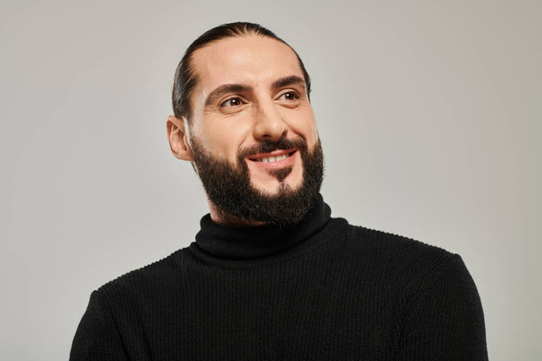Porträt eines glücklichen und gut aussehenden arabischen Mannes mit Bart, der in schwarzem Rollkragen vor grauem Hintergrund posiert - Foto, Bild