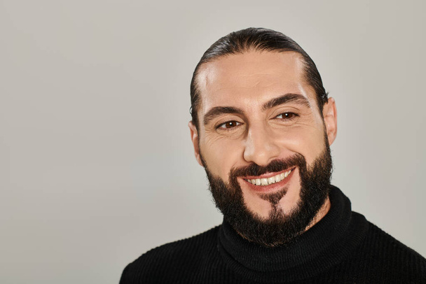 portrait de joyeux homme arabe avec barbe posant en col roulé noir sur fond gris - Photo, image