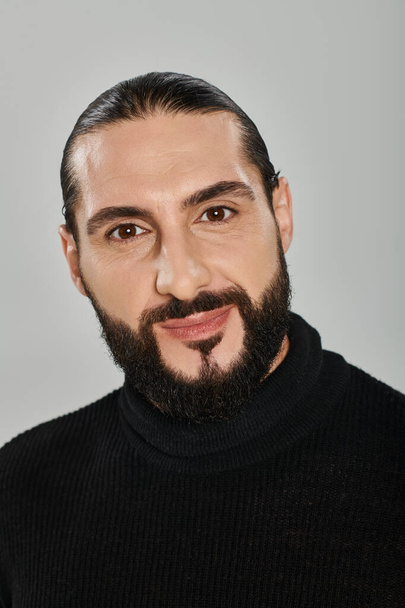 portrait of happy good looking arabic man with beard posing in turtleneck on grey backdrop - Photo, Image