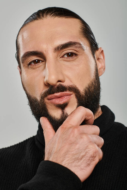 Porträt eines gut aussehenden arabischen Mannes in Rollkragen, der Bart berührt und vor grauem Hintergrund posiert - Foto, Bild