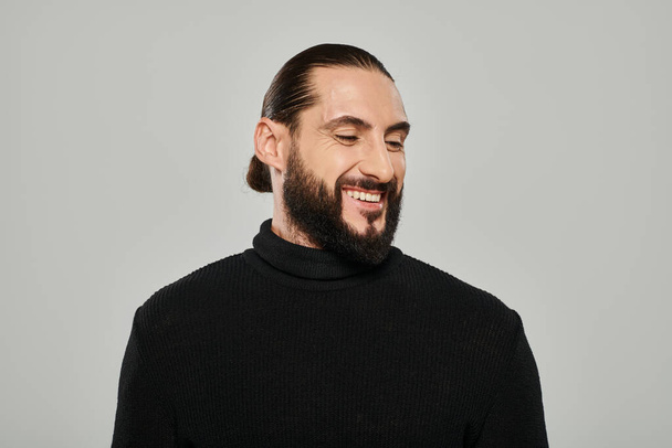 Porträt eines gut aussehenden arabischen Mannes mit Bart, der im Rollkragen posiert und vor grauem Hintergrund lächelt - Foto, Bild