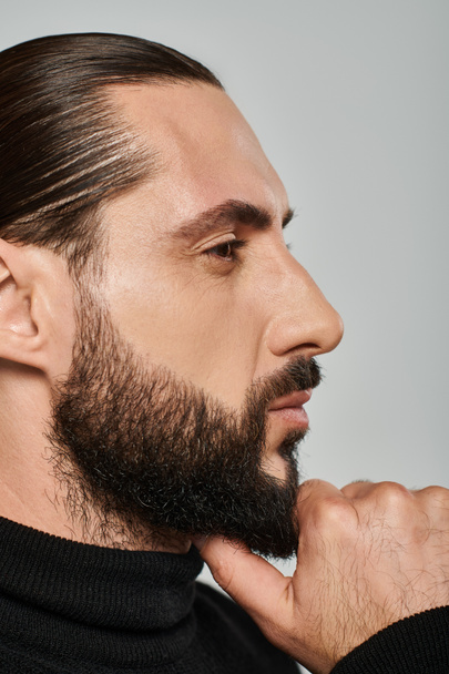 profile of good looking arabic man in turtleneck touching beard while thinking on grey backdrop - Photo, Image