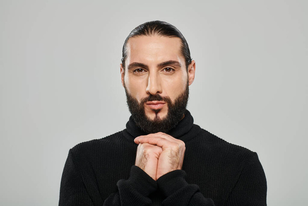 Porträt eines bärtigen arabischen Mannes mit schwarzem Rollkragen, der vor grauem Hintergrund in die Kamera blickt - Foto, Bild