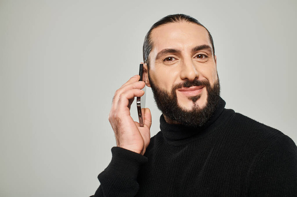 hombre árabe feliz con la barba sonriendo y tener llamada telefónica en el teléfono inteligente sobre fondo gris - Foto, Imagen