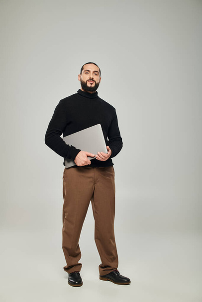 pohledný a vousatý arabský muž s úsměvem a stojící s notebookem na šedém pozadí, na volné noze - Fotografie, Obrázek