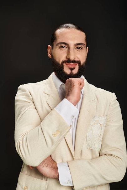 hombre árabe barbudo alegre en camisa blanca y chaqueta mirando a la cámara sobre fondo negro - Foto, imagen