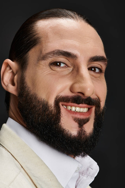 close up portrait of bearded and joyful arabic man in elegant attire posing on black backdrop - Photo, Image