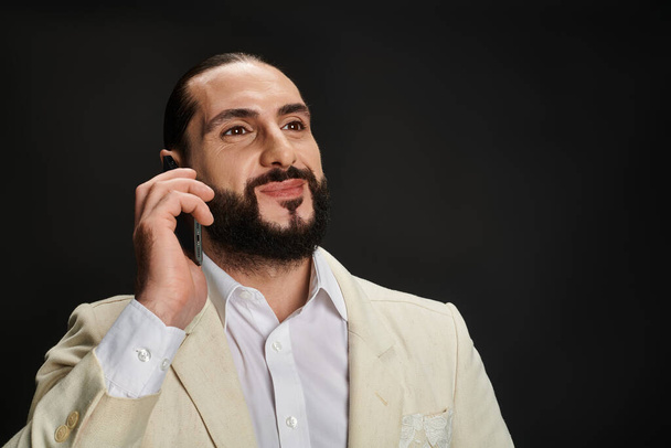 hombre árabe alegre y barbudo en camisa blanca y chaqueta hablando en smartphone sobre fondo negro - Foto, Imagen