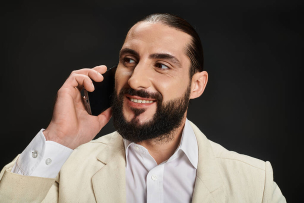 fröhlicher, bärtiger arabischer Mann in weißem Hemd und Blazer, der auf schwarzem Hintergrund mit dem Smartphone spricht - Foto, Bild