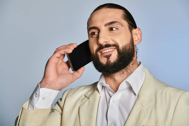 χαρούμενος γενειοφόρος αραβικός άνθρωπος σε κομψό επίσημη φορούν μιλάμε για smartphone σε γκρι φόντο, τηλεφώνημα - Φωτογραφία, εικόνα