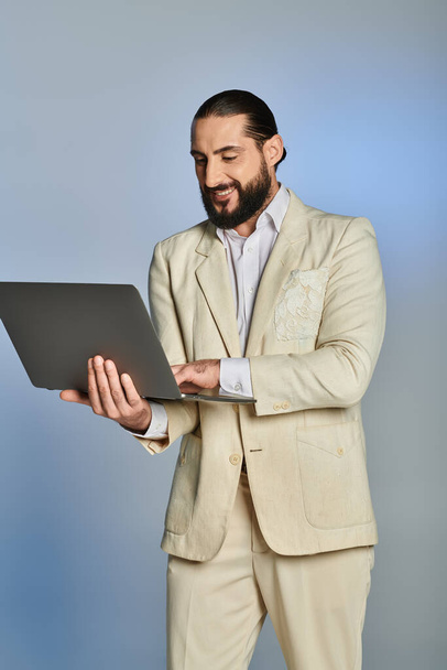 hombre árabe barbudo feliz en ropa formal elegante usando el ordenador portátil sobre fondo gris, era digital - Foto, Imagen