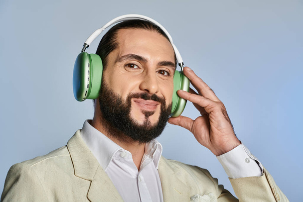 χαρούμενος γενειοφόρος άνδρας σε κομψό επίσημη φθορά ακούγοντας μουσική σε ασύρματα ακουστικά σε γκρι φόντο - Φωτογραφία, εικόνα