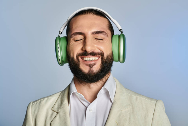 ευτυχισμένος γενειοφόρος άνδρας σε κομψό επίσημη φθορά ακούγοντας μουσική σε ασύρματα ακουστικά σε γκρι, κλειστά μάτια - Φωτογραφία, εικόνα