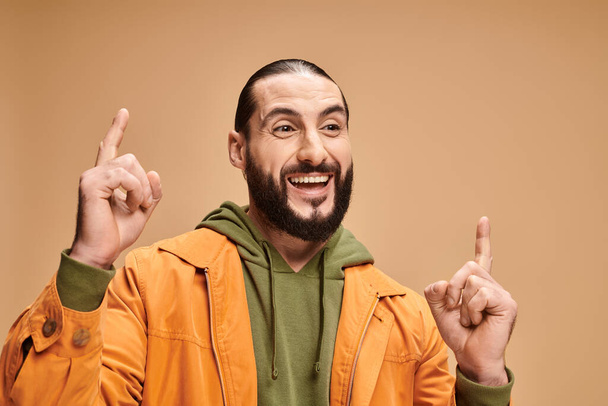 portrait of positive and bearded arabic man in casual attire pointing up on beige background - Photo, Image