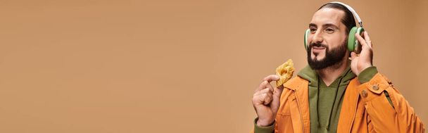 cheerful man in headphones holding honey baklava on beige background, middle eastern dessert banner - Photo, Image