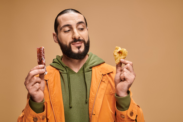 hombre barbudo alegre sonriendo y sosteniendo baklava y cevizli sucuk sobre fondo beige, delicias turcas - Foto, Imagen