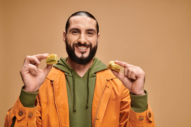 happy bearded man holding two different kinds of baklava on beige backdrop, turkish delights - Фото, изображение