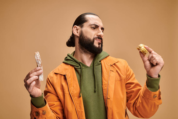 confuso hombre barbudo árabe sosteniendo baklava y cevizli sucuk sobre fondo beige, delicias turcas - Foto, imagen