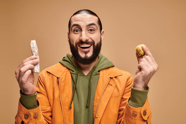 hombre barbudo árabe excitado sosteniendo baklava y cevizli sucuk sobre fondo beige, delicias turcas - Foto, imagen