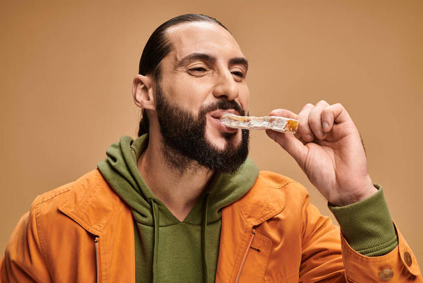 cheerful and bearded arabic man eating delicious cevizli sucuk on beige backdrop, turkish delights - Photo, Image