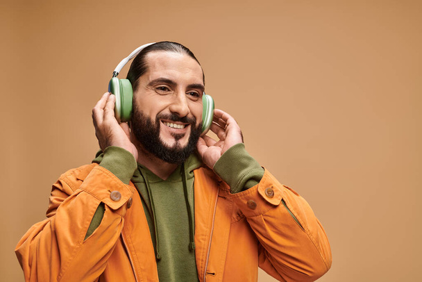 homme du Moyen-Orient gai avec barbe écouter de la musique dans des écouteurs sans fil sur fond beige - Photo, image