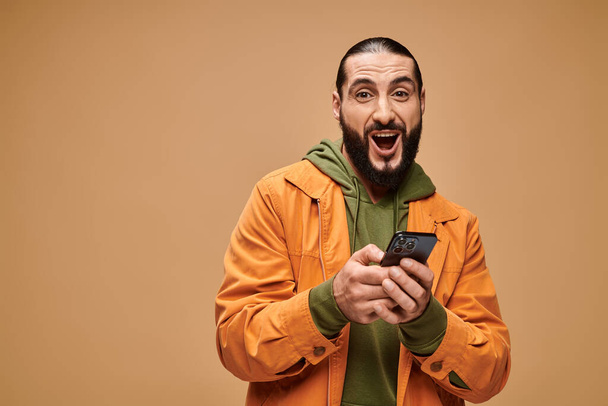 felice uomo mediorientale con barba utilizzando smartphone su sfondo beige, era digitale - Foto, immagini