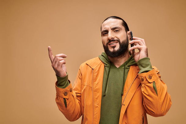 емоційний східний чоловік з бородою розмовляє на смартфоні на бежевому фоні, банер - Фото, зображення